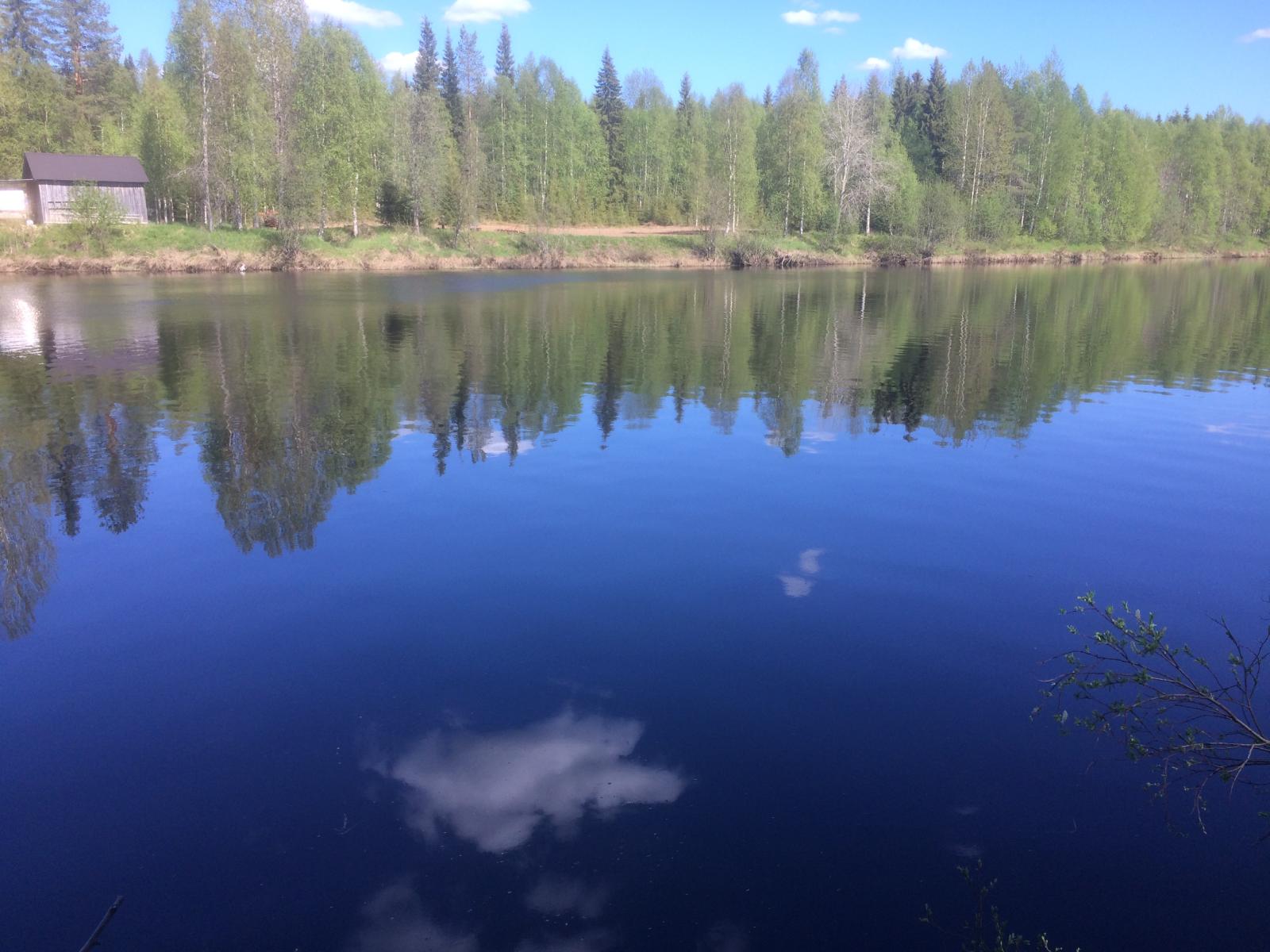 Vastarannan metsä ja lähes pilvetön taivas heijastuvat joen tyyneen pintaan. Rannassa on rakennus.