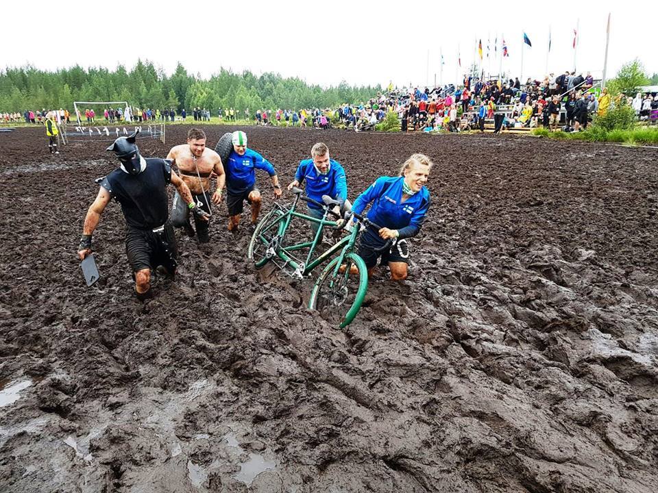 Suojuoksutapahtumassa viisi kilpailijaa ylittää mutaista suota taluttaen tandem-pyörää.