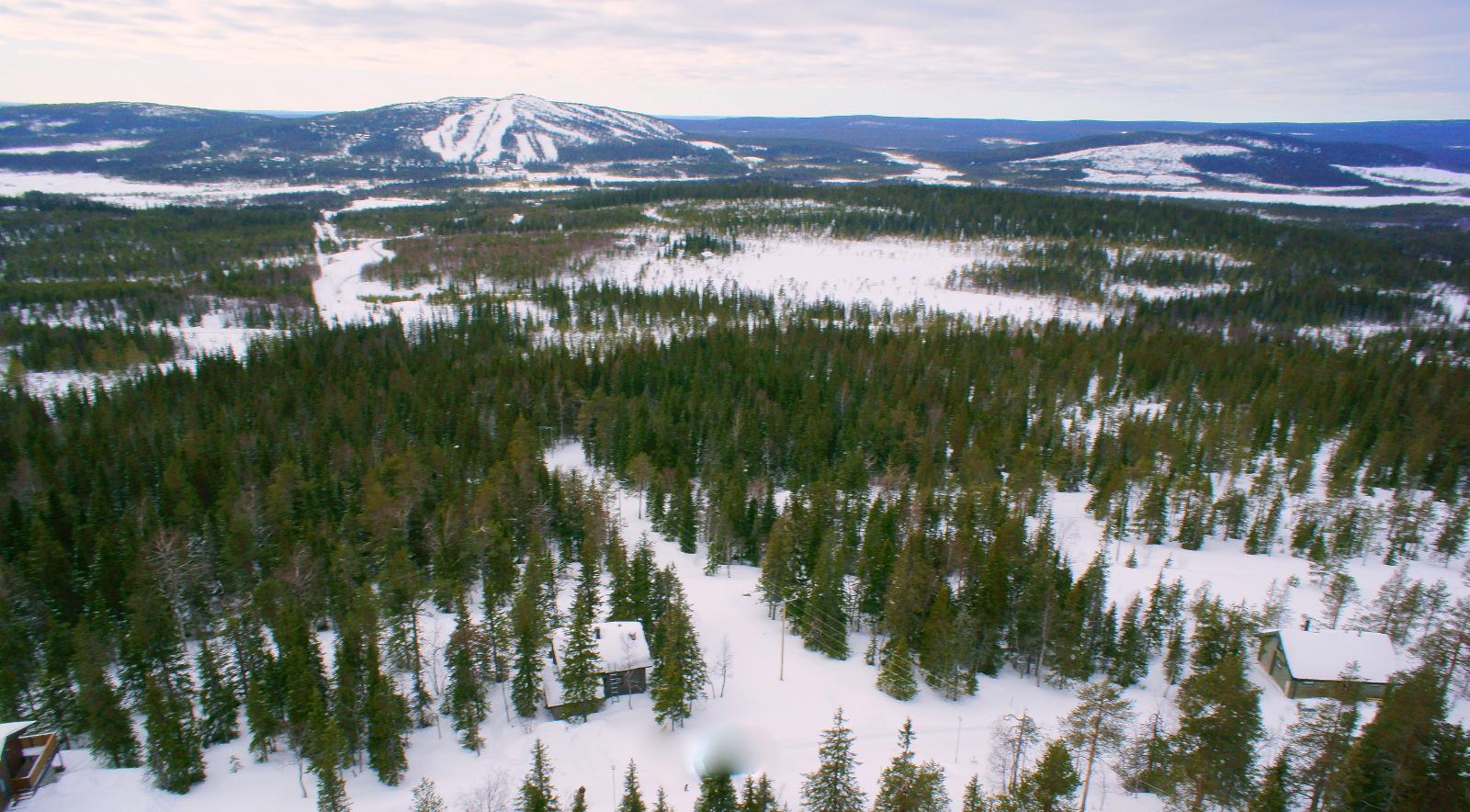 Lumipeitteisessä metsässä on mökkejä. Taustalla kohoaa tunturimaisema. Ilmakuva.