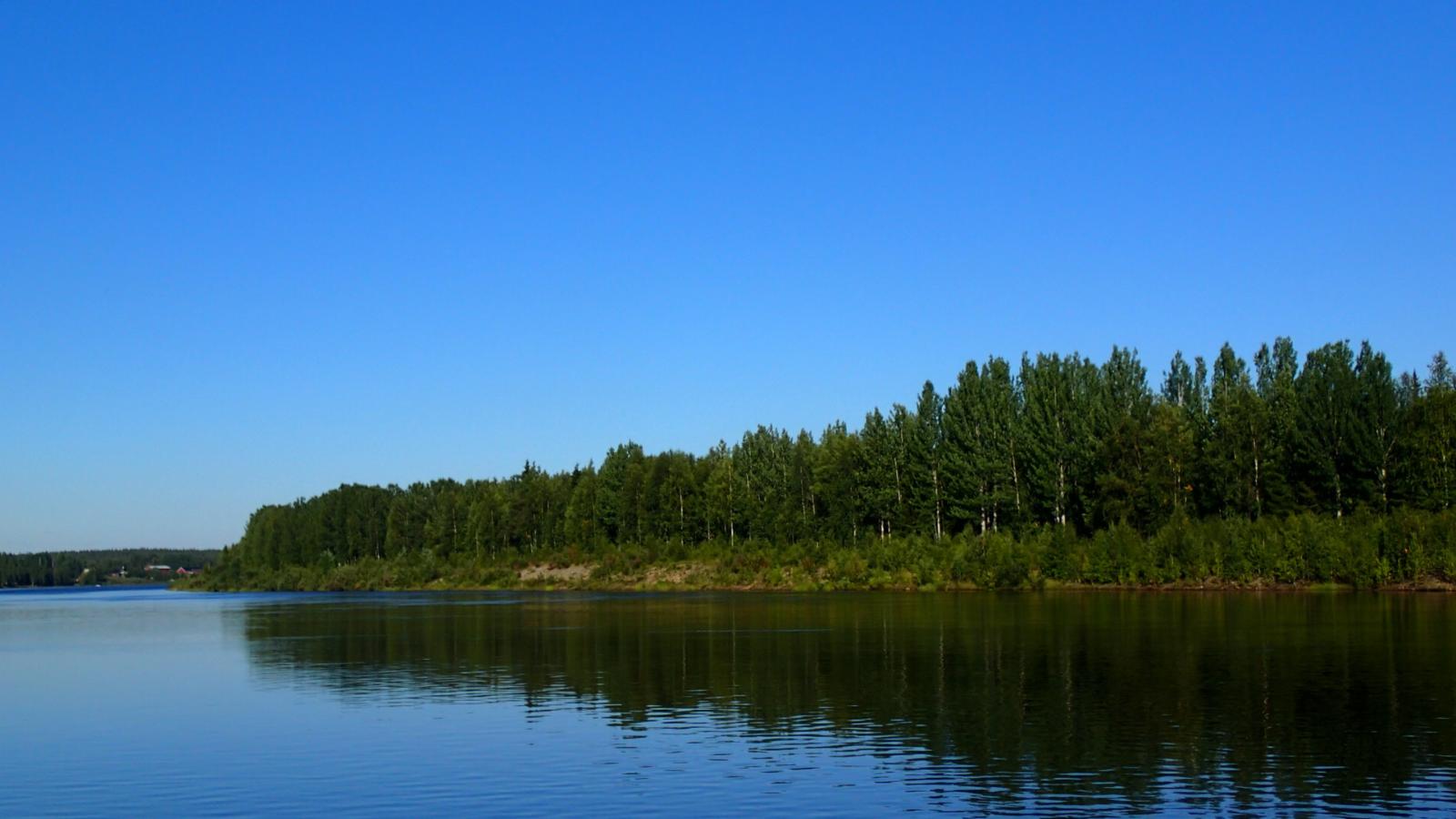 Joen vastarannan metsä heijastuu joen pintaan pilvettömänä päivänä. Horisontissa häämöttää rakennus.