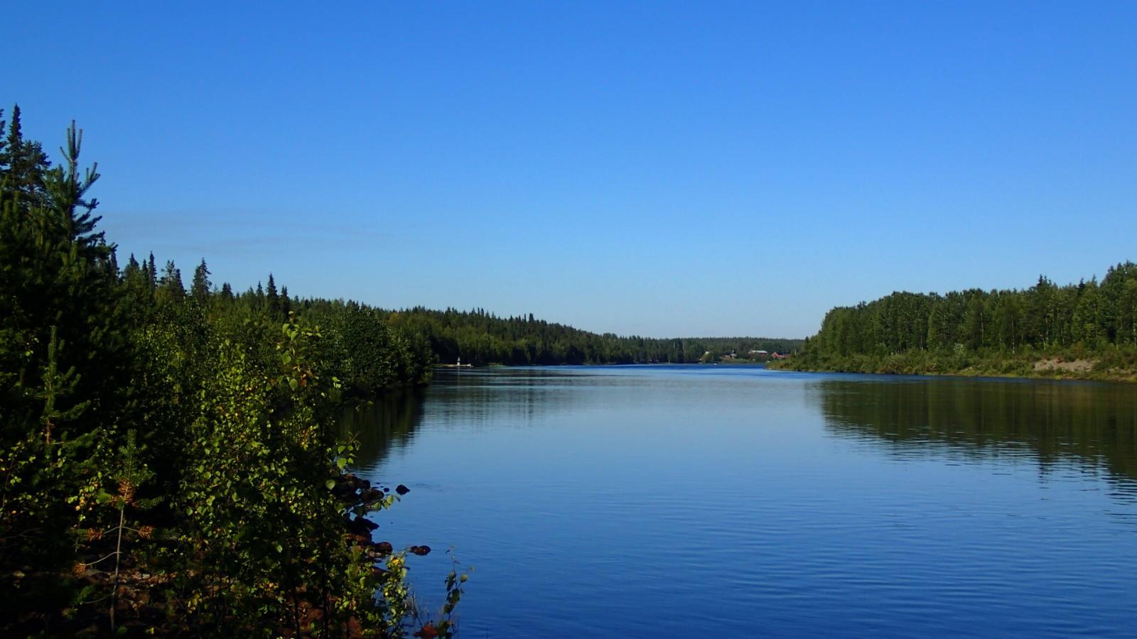 Kitinen virtaa metsien halki. Horisontissa joen rannalla on rakennuksia.