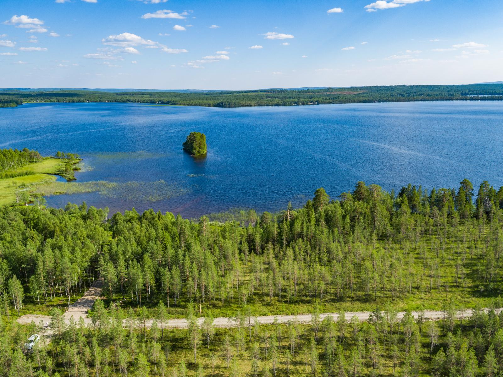 Kesämaisemassa metsä reunustaa järveä, jossa on saari. Etualan metsässä kulkee tie. Ilmakuva.