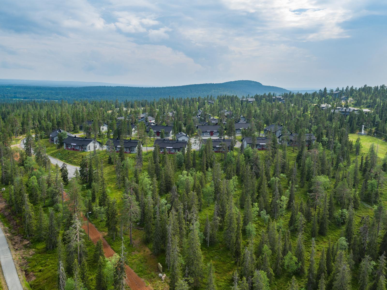 Mökkialuetta ympäröi kuusivaltainen metsä. Horisontissa kohoaa vaaramaisema. Ilmakuva.