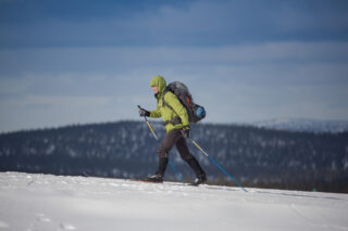 Hiihtäjä kulkee puuttomalla tunturin rinteellä reppu selässä. Taka-alalla kohoaa tunturimaisema.