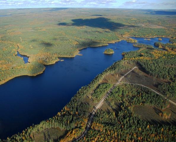 Pitkulaista järveä reunustaa horisonttiin ulottuva metsä. Rannan lähellä kulkee tie. Ilmakuva.