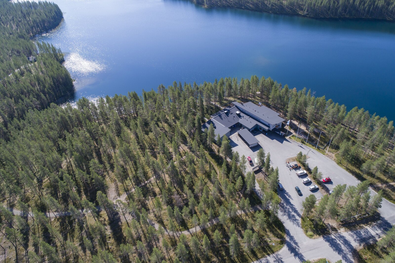 Metsä reunustaa järvimaisemaa. Järven rannalla luontokeskuksen rakennus. Ilmakuva.