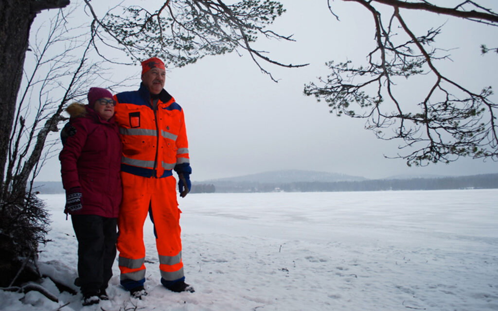 Pariskunta seisoo ison puun alla järven rannalla. Järvi on jäässä.