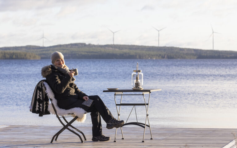 Nainen istuu lämpimissä vaatteissa laiturilla juomassa kahvia. Hän on iloinen.