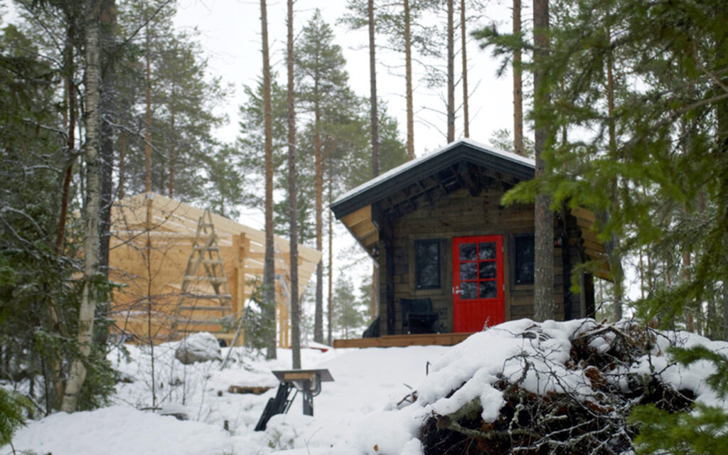 Valmis sauna, jossa on punainen tupa. Sen taustalle nousee varsinainen mökki.e