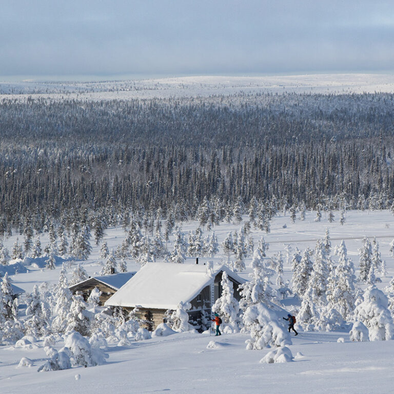 Talvinen tunturimaisema. Kaksi henkilöä on hiihtämässä kuvassa näkyvien mökkien edessä.