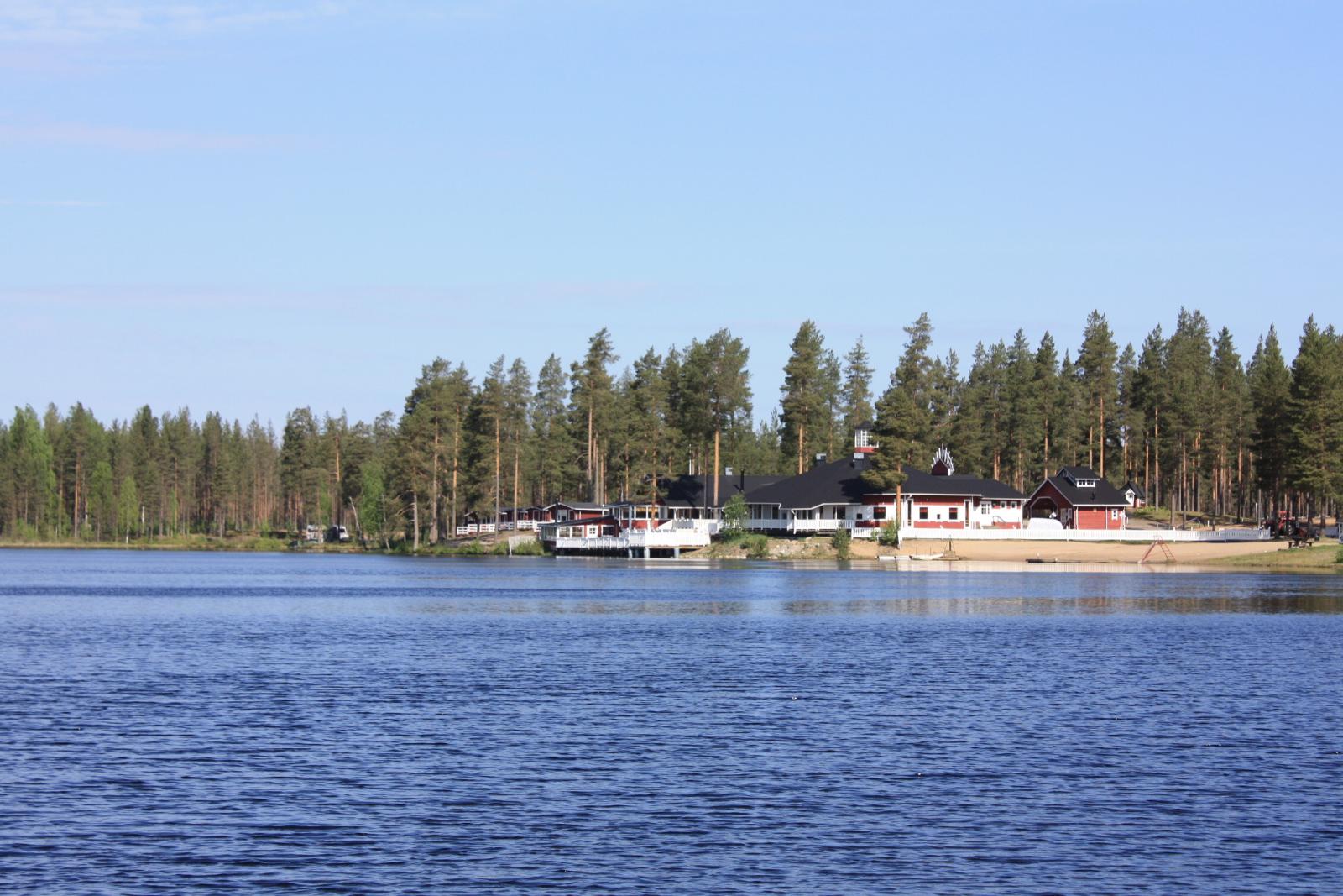 Kesäisessä järvimaisemassa vastarannalla on leirintäalueen rakennuksia. Veden pinta väreilee hieman.