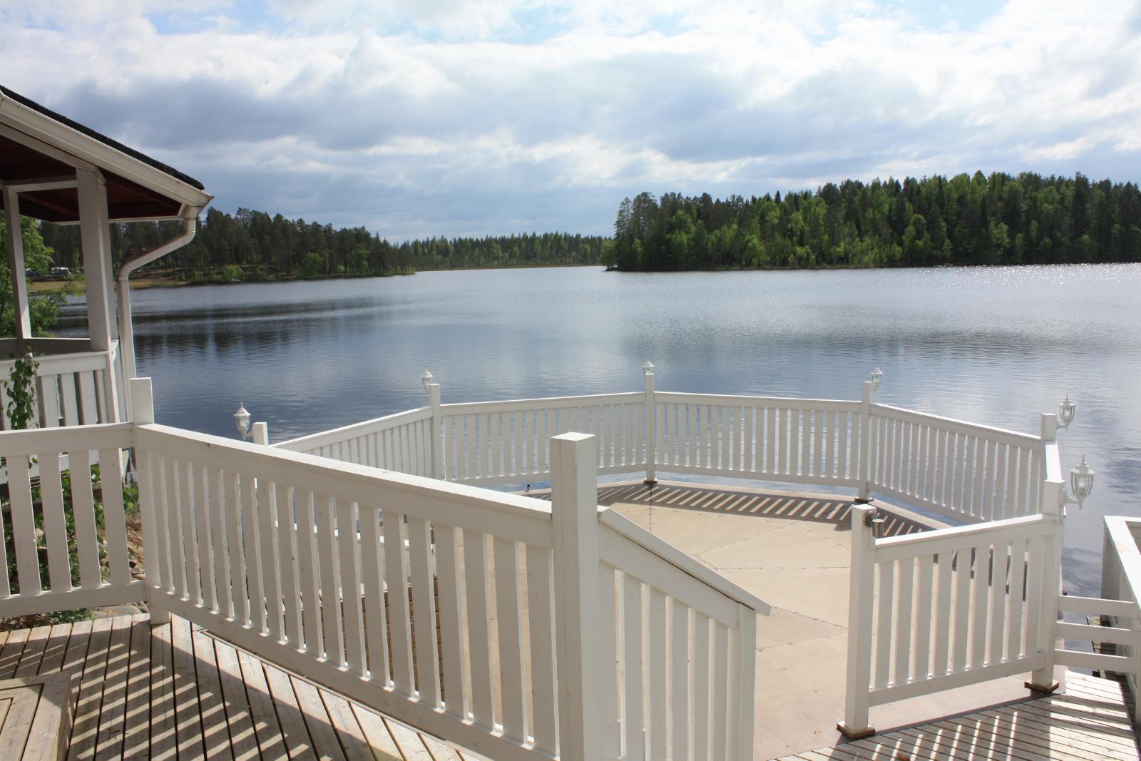 Aurinkoinen pyöreä terassi rakennuksen yhteydessä järven rannalla. Taivaalla poutapilviä.