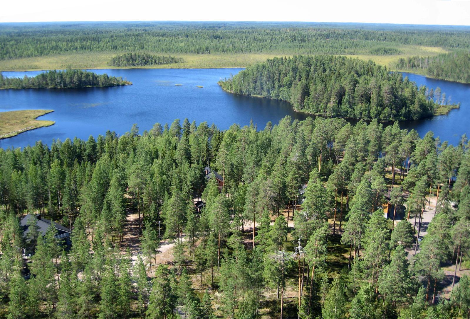 Metsät ja suot ympäröivät järveä, jossa on kaksi saarta. Puiden välistä erottuu mökkejä. Ilmakuva.