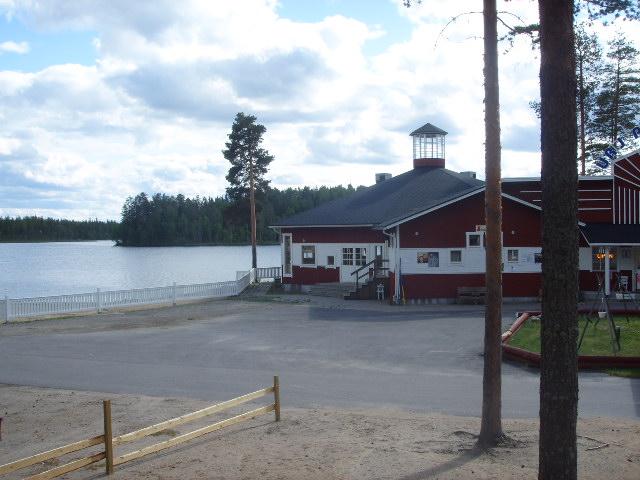 Urjanlinnan päärakennus piha-alueineen järven rannalla. Rannassa on korkea mänty.