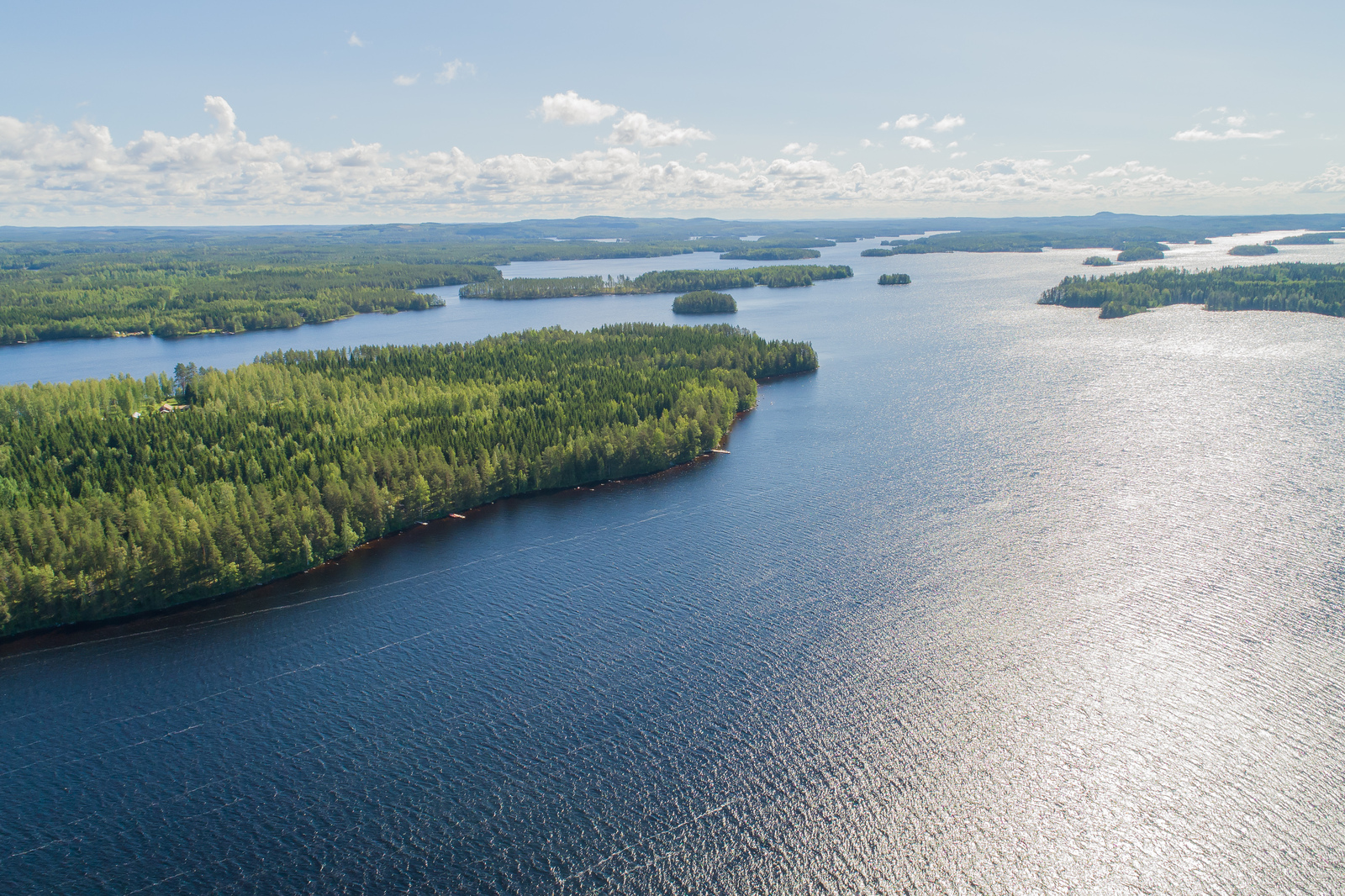 Metsäinen saari järveltä päin katsottuna, valkoisia pilviä aurinkoisella taivaalla.