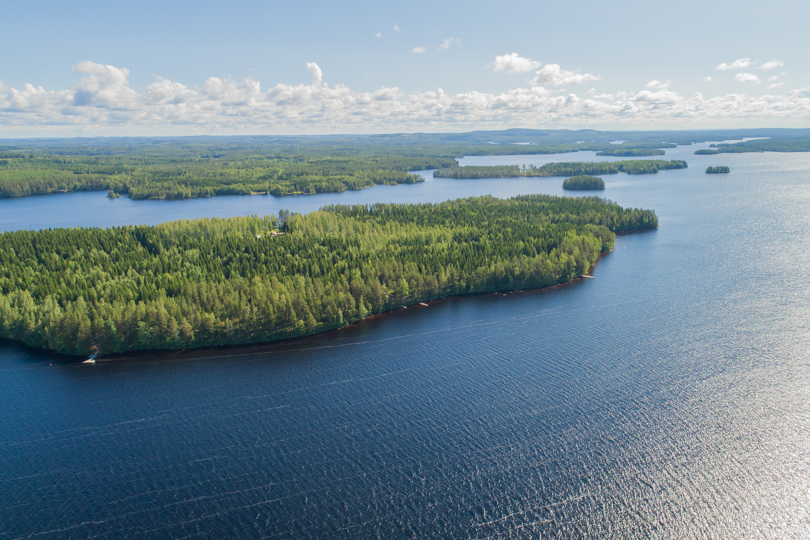 Metsäinen saari järveltä päin katsottuna, valkoisia pilviä aurinkoisella taivaalla.