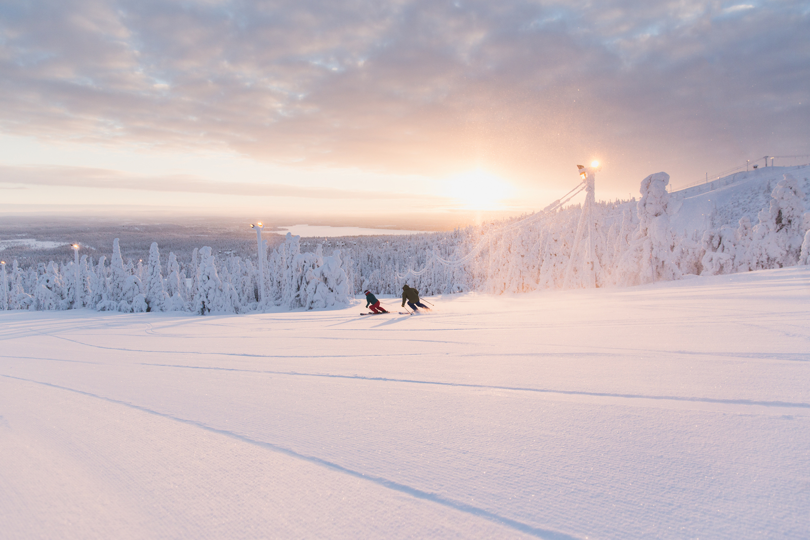 Kaksi laskettelijaa rinteessä. Taustalla tykkypuita ja auringonpaiste.