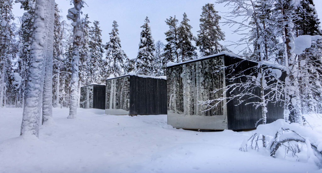 Kolme majoitusrakennusta talvisen metsän keskellä. Rakennusten pääty on lasipintaa.