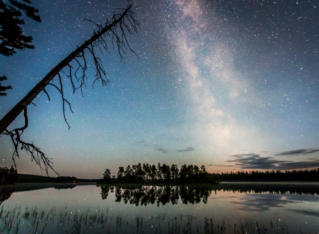 Syksyinen yötaivas erämaajärven rannalla. Kirkas tähtitaivas heijastuu järven pinnassa.