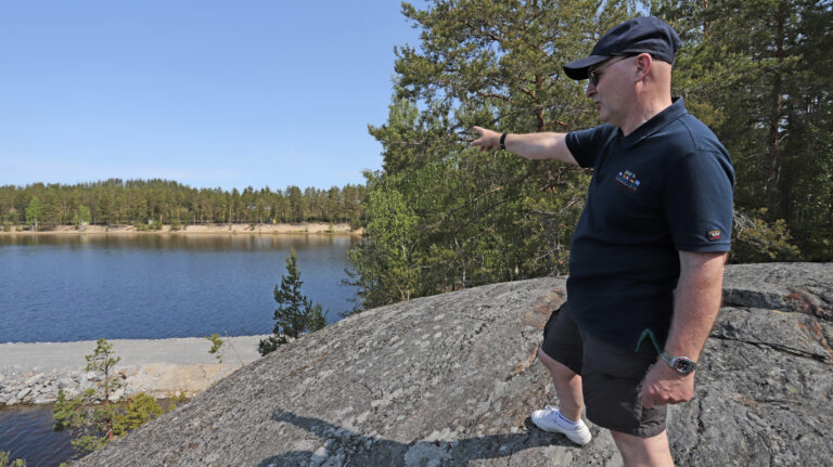 Mies seisoo kalliolla ja osoittaa lahden takana näkyvää rantaa.