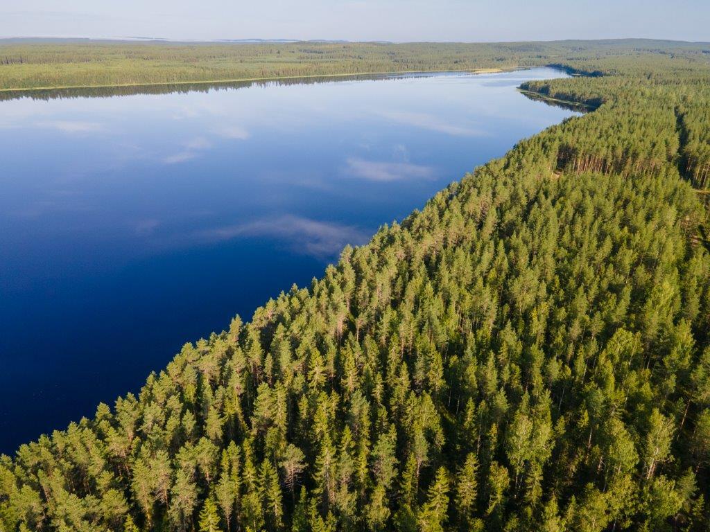 Ilmakuvassa sininen tyyni järvi josta heijastuu valkoisia pilviä, oikealla mäntymetsää.