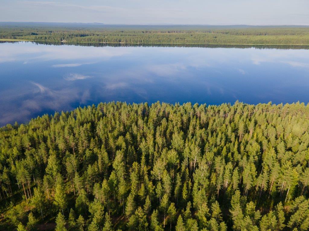 Ilmakuvassa sininen tyyni järvi josta heijastuu valkoisia pilviä, etualalla metsää.