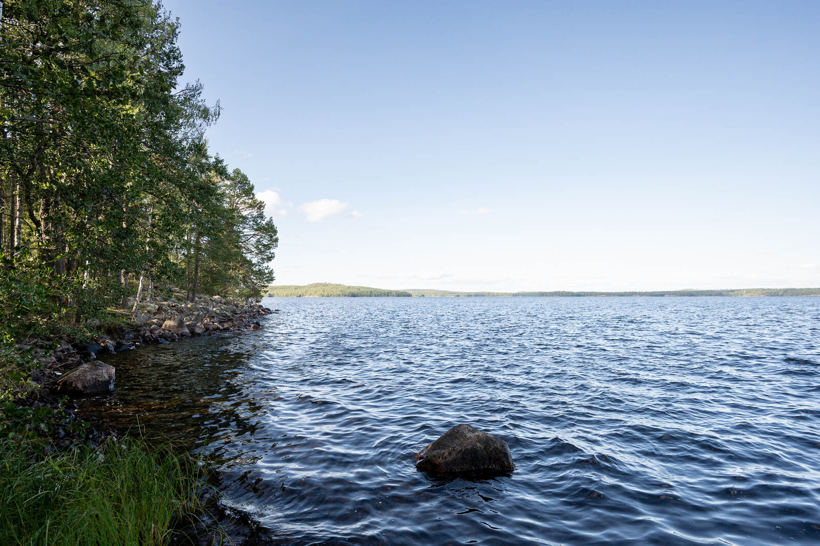 Näkymä ulapalle tontin rannasta, vasemmalla puustoa rannalla. Edessä vedessä isohko kivi.