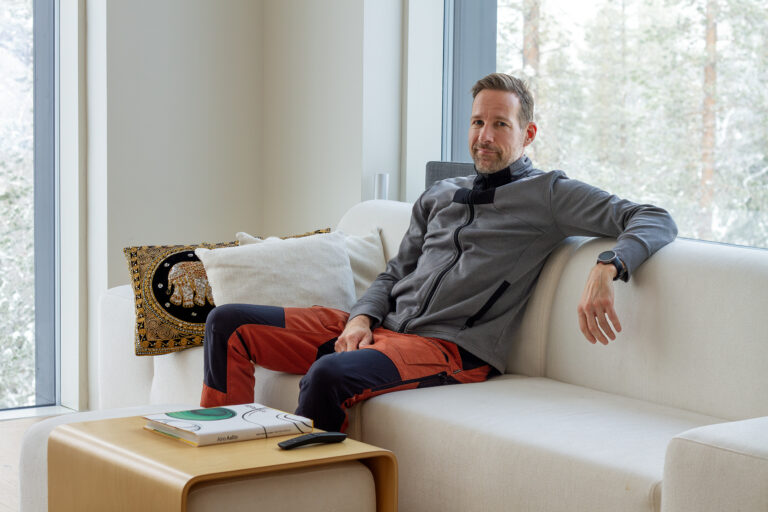 Mies istuu asunnossa vaalealla sohvalla. Ikkunoista näkyy ympäröivä talvinen metsämaisema.